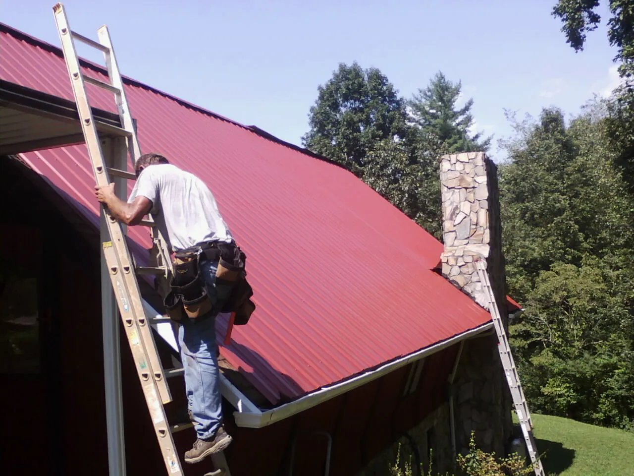 Roofer on a ladder beside a red metal roof and stone chimney — Soffit & Fascia Repair by Roofing Brothers in Wareham